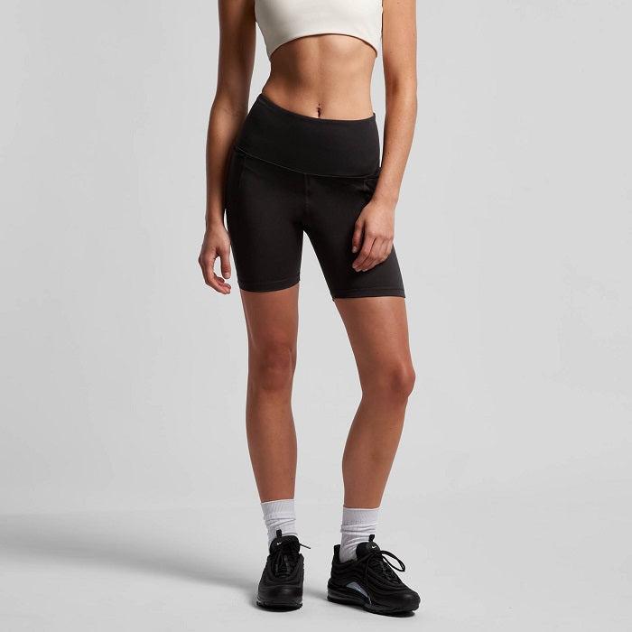 A female model wearing black active bike shorts with side pockets, paired with a white top and black sneakers.