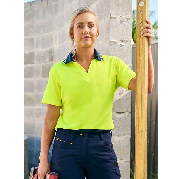 Person wearing a bright yellow safety shirt and navy pants holding a long wooden tool against a stone wall.