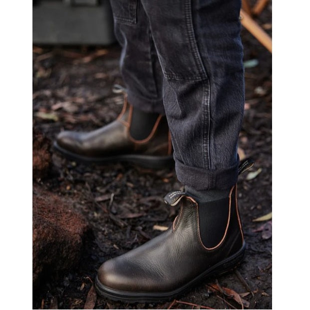 Dark brown leather boots worn with dark jeans on a natural ground surface.