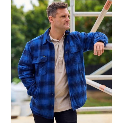 Man wearing a blue plaid shirt leaning against a railing outdoors.