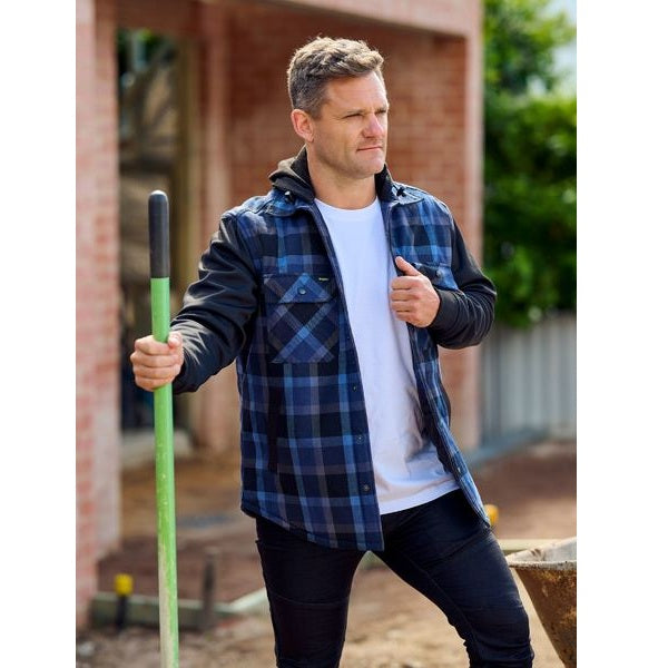 Man wearing a blue plaid shirt over a white t-shirt, holding a green tool outdoors.