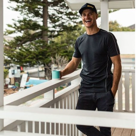 Man in black athletic wear holding a blue cup on a balcony with trees and a house in the background