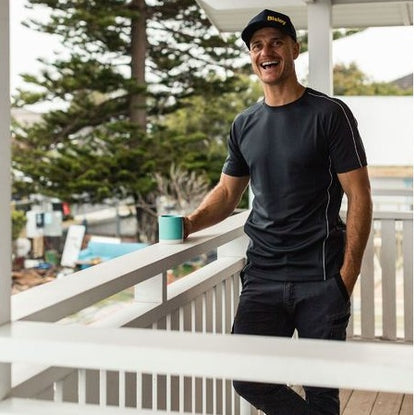 Man in black athletic wear holding a blue cup on a balcony with trees and a house in the background