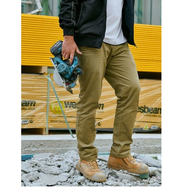 Person wearing tan work pants and brown boots holding a power tool on a construction site.