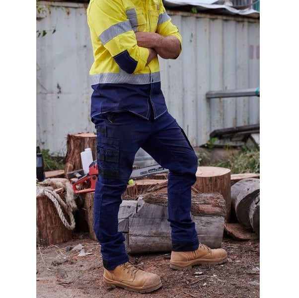Person wearing a yellow safety jacket and blue work pants in an outdoor setting with logs and a metal container.
