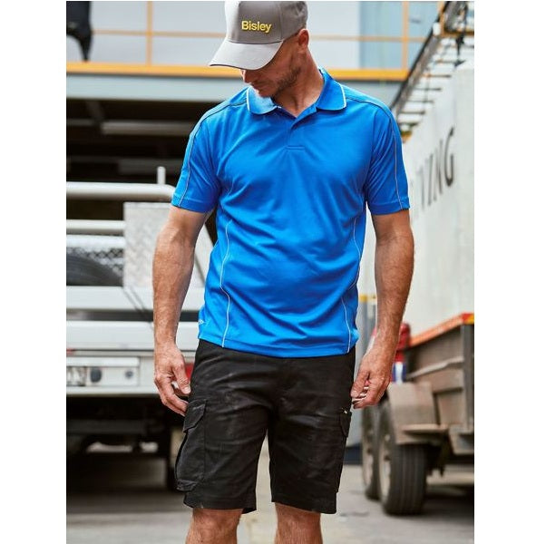 Man wearing a blue shirt and black shorts with a visible brand logo, standing in front of trucks.