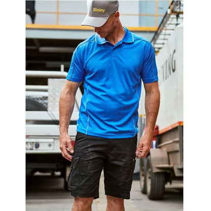 Man wearing a blue shirt and black shorts with a visible brand logo, standing in front of trucks.