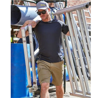 Man wearing a navy shirt and beige shorts leaning against metal pipes with a blue barrel in the background.