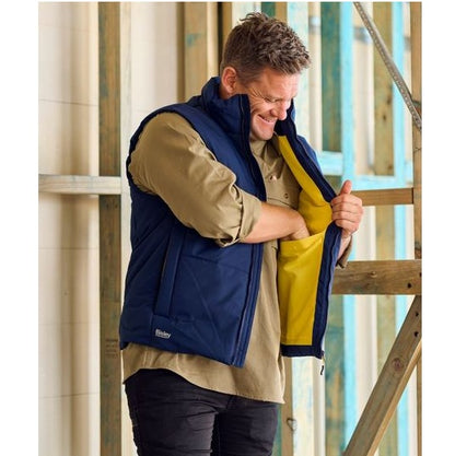 Man wearing a blue and yellow jacket with a visible brand logo, standing in front of wooden scaffolding.