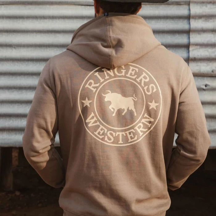 A man from behind wearing a tan hoodie with a large cream circular 'Ringers Western' bull logo graphic on the back, standing in front of a farm shed.