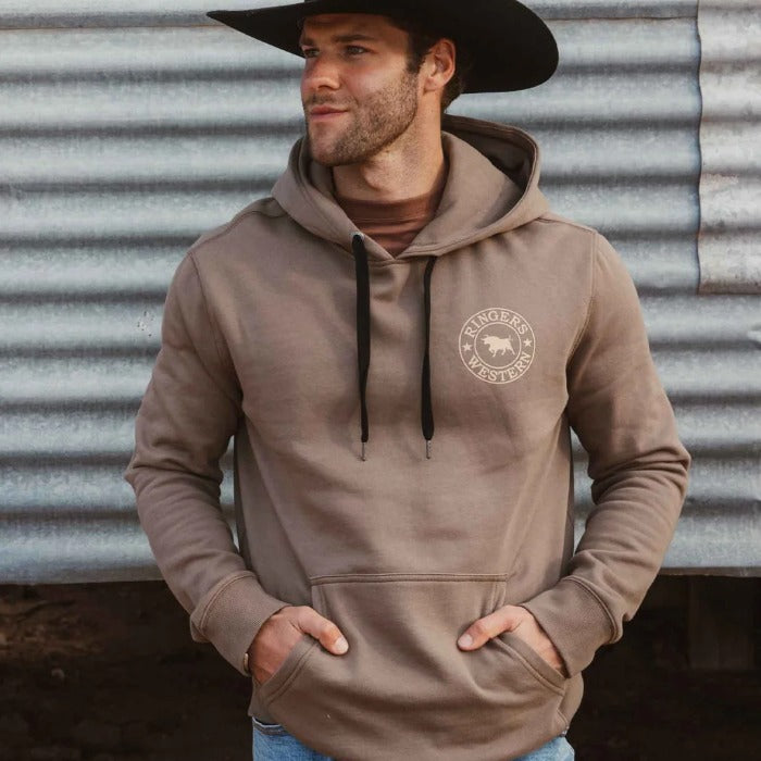 A man wearing a taupe-brown pullover hoodie with a circular 'Ringers Western' bull logo on the left chest, paired with a black cowboy hat in Australia outback